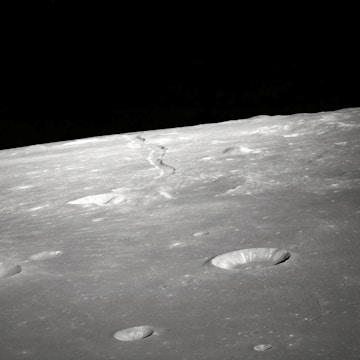 A barren, grey lunar landscape with multiple craters of varying sizes scattered across the surface. A distinct meandering line runs toward the horizon, possibly indicating a ridge or fault line. The dark sky above provides stark contrast to the illuminated surface.