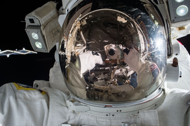An astronaut in a spacesuit with a reflective helmet visor is shown. The reflection in the visor captures parts of a spacecraft, the astronaut's gloved hands, and a bright light source, possibly from the sun. The backdrop is the dark expanse of space.
