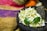 A collection of white flowers with green leaves rests in a rough dark stone molcajete. Behind them, a colorful woven textile with red, yellow, and purple hues is draped. Oranges and books partially obscure the background.
