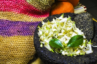 A collection of white flowers with green leaves rests in a rough dark stone molcajete. Behind them, a colorful woven textile with red, yellow, and purple hues is draped. Oranges and books partially obscure the background.