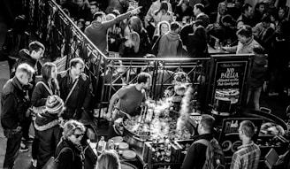 A crowded market stall with people gathered around a large pan where food is being cooked. Steam rises from the pan while various market-goers, dressed in winter clothing, interact and observe. A sign nearby indicates rules about paella in the courtyard.