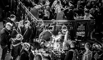 A crowded market stall with people gathered around a large pan where food is being cooked. Steam rises from the pan while various market-goers, dressed in winter clothing, interact and observe. A sign nearby indicates rules about paella in the courtyard.