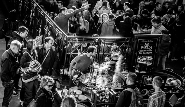 A crowded market stall with people gathered around a large pan where food is being cooked. Steam rises from the pan while various market-goers, dressed in winter clothing, interact and observe. A sign nearby indicates rules about paella in the courtyard.