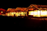 A series of greenhouses are illuminated against the night sky, casting a warm, glowing light through their glass panes. The structures are side by side, with distinct, angular roofs, and surrounded by darkness, which emphasizes the bright light within.