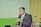 A person in formal attire is speaking into a microphone. The individual is wearing a suit and tie, standing behind a podium. The background features a green and white wall with a yellow curtain to the side, and decorative flowers on the right.