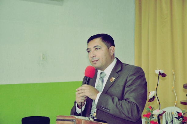 A person in formal attire is speaking into a microphone. The individual is wearing a suit and tie, standing behind a podium. The background features a green and white wall with a yellow curtain to the side, and decorative flowers on the right.