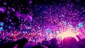 An energetic scene with vibrant colors dominating the atmosphere. There are numerous bubbles or foam particles floating in the air, illuminated by bright pink, purple, and blue lights. Silhouettes of people with raised hands suggest a festive, lively event, such as a concert or party.
