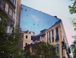 A building's wall features a large mural that depicts urban architecture with detailed facades and windows. Birds are also painted, giving an illusion of depth and continuation. In the foreground, lush greenery and a vintage streetlamp add a natural element to the scene.