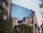 A building's wall features a large mural that depicts urban architecture with detailed facades and windows. Birds are also painted, giving an illusion of depth and continuation. In the foreground, lush greenery and a vintage streetlamp add a natural element to the scene.