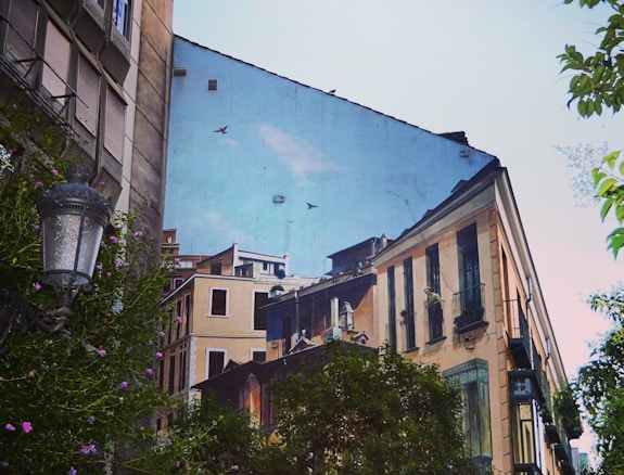 A building's wall features a large mural that depicts urban architecture with detailed facades and windows. Birds are also painted, giving an illusion of depth and continuation. In the foreground, lush greenery and a vintage streetlamp add a natural element to the scene.