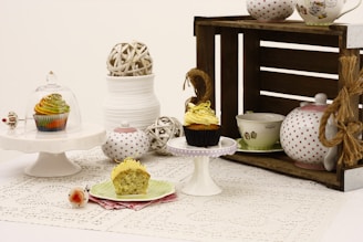 A vintage-style table setting featuring cupcakes on small cake stands. One cupcake is under a glass dome, while another is partially eaten on a green plate with a pink napkin. A rustic wooden crate holds polka dot and decorated teapots and cups. Various decorative balls and a stack of white bowls add to the composition.