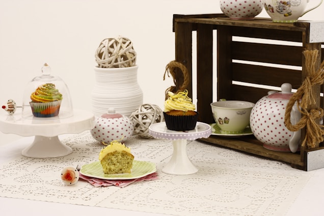 A vintage-style table setting featuring cupcakes on small cake stands. One cupcake is under a glass dome, while another is partially eaten on a green plate with a pink napkin. A rustic wooden crate holds polka dot and decorated teapots and cups. Various decorative balls and a stack of white bowls add to the composition.