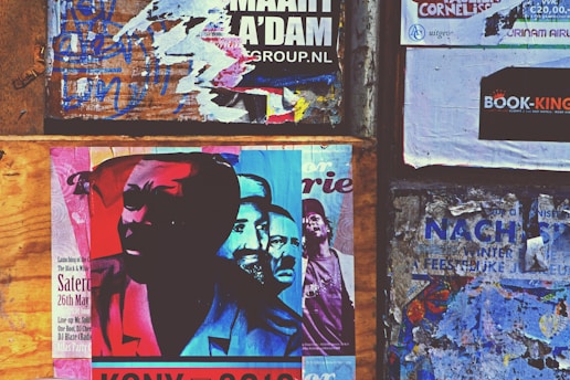 A collage of posters and advertisements pasted on a wooden and concrete wall. The main poster features an abstract, colorful image of multiple faces with red, blue, and cyan hues. Surrounding the central poster are various torn and overlapping flyers with visible text and graphics in different languages, adding a sense of urban chaos.