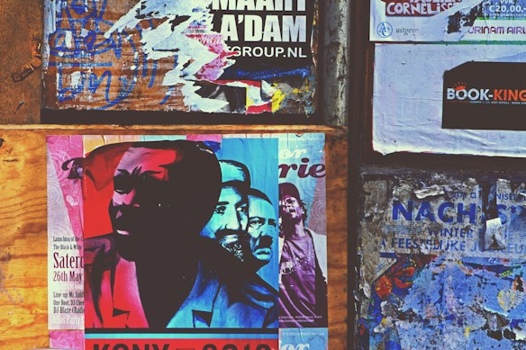A collage of posters and advertisements pasted on a wooden and concrete wall. The main poster features an abstract, colorful image of multiple faces with red, blue, and cyan hues. Surrounding the central poster are various torn and overlapping flyers with visible text and graphics in different languages, adding a sense of urban chaos.
