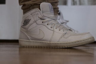 A close-up of a white sneaker with visible stitching and a logo on the side. The shoe is worn, showing slight signs of use, and is positioned on a wooden floor. The sneaker is laced up and paired with light brown pants.