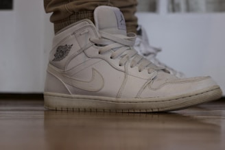 A close-up of a white sneaker with visible stitching and a logo on the side. The shoe is worn, showing slight signs of use, and is positioned on a wooden floor. The sneaker is laced up and paired with light brown pants.