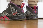 A pair of stylish sneakers featuring a black and speckled design with red accents. The sneakers include prominent branding and are worn by someone standing on a wooden floor.