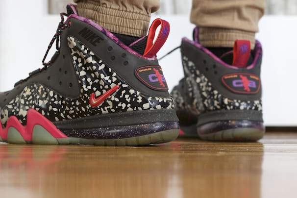 A pair of stylish sneakers featuring a black and speckled design with red accents. The sneakers include prominent branding and are worn by someone standing on a wooden floor.