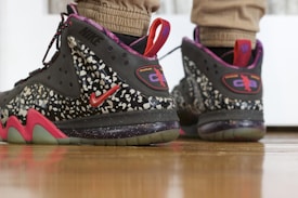 A pair of stylish sneakers featuring a black and speckled design with red accents. The sneakers include prominent branding and are worn by someone standing on a wooden floor.