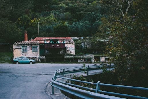 An abandoned building with a worn-out facade sits near a winding asphalt road. A small, turquoise car is parked on the left side close to the building. The building has a chimney and a tiled roof, with overgrown foliage and trees surrounding it. The scene is set against a backdrop of dense forested hills, and there are guardrails lining the curved road.