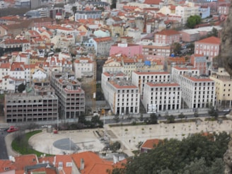 A densely packed urban landscape with a mix of modern and traditional architecture. The buildings predominantly feature red-tiled roofs and are closely arranged. Several multi-story structures are under construction or appear incomplete, adding an element of urban development. A large open space or plaza is visible in the foreground, with trees and pathways. The background showcases a variety of architectural designs.