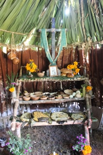An altar is set up inside a structure with palm leaves covering the top. The altar includes a large wooden cross hanging at the center with a green woven cloth draped over it. It is adorned with various offerings, including bowls of food, fruits, flowers, and bread. Brightly colored marigold flowers are also placed around the altar, and there are small plants at the base.