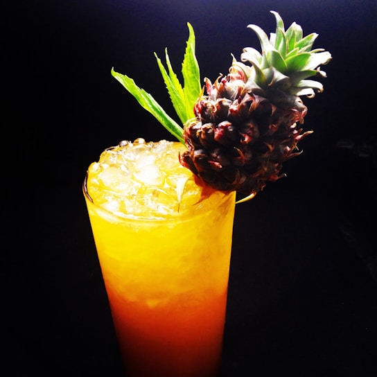 A tall glass filled with a vibrant yellow-orange beverage topped with crushed ice. Garnished with a small, whole pineapple and a few green leaves, set against a dark background.