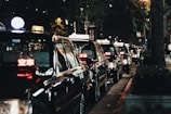 A line of black taxis parked along a dimly lit street at night, illuminated by streetlights. The taxis have lit signs on top with glowing red lettering, indicating they are available for passengers. Trees and urban elements are visible in the background, contributing to a city atmosphere.