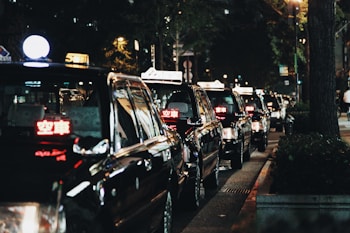 A line of black taxis parked along a dimly lit street at night, illuminated by streetlights. The taxis have lit signs on top with glowing red lettering, indicating they are available for passengers. Trees and urban elements are visible in the background, contributing to a city atmosphere.