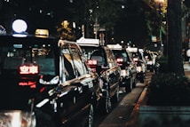 A line of black taxis parked along a dimly lit street at night, illuminated by streetlights. The taxis have lit signs on top with glowing red lettering, indicating they are available for passengers. Trees and urban elements are visible in the background, contributing to a city atmosphere.