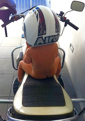 A young child sitting on a motorcycle seat, wearing an oversized helmet that covers their head. The child is facing away, with their feet resting on the motorcycle frame. The setting appears to be a garage or enclosed space with a tiled floor and white walls.