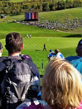 A crowd gathers around a golf course where a golfer, dressed in dark clothing, is seen walking on the green towards a flag. An electronic scoreboard in the background displays scores, indicating a professional golf event. Surrounding the scoreboard is a seated crowd on a grassy hill, enjoying the sunny day.