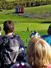 A crowd gathers around a golf course where a golfer, dressed in dark clothing, is seen walking on the green towards a flag. An electronic scoreboard in the background displays scores, indicating a professional golf event. Surrounding the scoreboard is a seated crowd on a grassy hill, enjoying the sunny day.