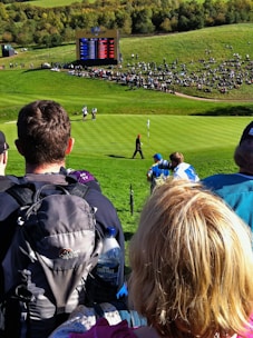 A crowd gathers around a golf course where a golfer, dressed in dark clothing, is seen walking on the green towards a flag. An electronic scoreboard in the background displays scores, indicating a professional golf event. Surrounding the scoreboard is a seated crowd on a grassy hill, enjoying the sunny day.