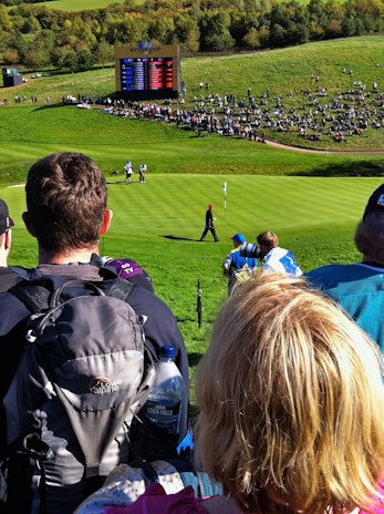 A crowd gathers around a golf course where a golfer, dressed in dark clothing, is seen walking on the green towards a flag. An electronic scoreboard in the background displays scores, indicating a professional golf event. Surrounding the scoreboard is a seated crowd on a grassy hill, enjoying the sunny day.