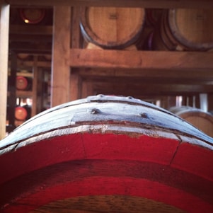 A close-up of a wooden barrel with red accents in a dimly lit storage room. Other blurred barrels are visible in the background, stacked on wooden shelves.