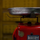A metal tray is attached to the top of a red base that appears to be part of a weighing scale. The metal tray is worn and has visible scratches and marks. There is a label with some text and numbers affixed to the red base. The background is yellow and slightly out of focus.