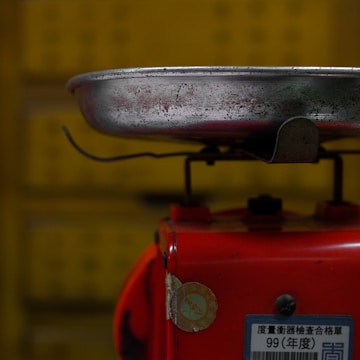 A metal tray is attached to the top of a red base that appears to be part of a weighing scale. The metal tray is worn and has visible scratches and marks. There is a label with some text and numbers affixed to the red base. The background is yellow and slightly out of focus.