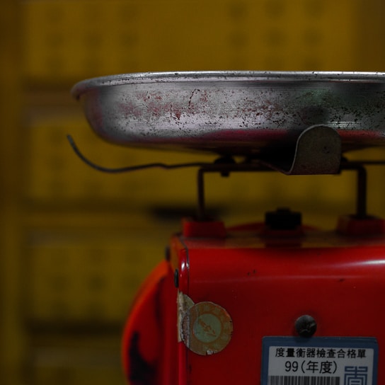 A metal tray is attached to the top of a red base that appears to be part of a weighing scale. The metal tray is worn and has visible scratches and marks. There is a label with some text and numbers affixed to the red base. The background is yellow and slightly out of focus.