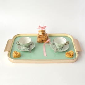 A pastel-colored tray holds two elegantly designed teacups and saucers, each accompanied by a small spoon. At the center of the tray, a bag filled with cookies tied with a pink ribbon is prominently placed. Heart-shaped cookies are placed beside each cup, adding a charming touch.