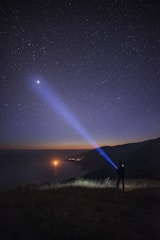 A person stands on a grassy hill, pointing a blue flashlight beam towards the sky in a starry night scene. The landscape below shows a coastline with water reflecting lights, possibly from distant boats or other sources. The horizon displays a subtle gradient of colors as the evening transitions into night.