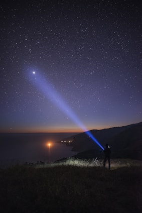 A person stands on a grassy hill, pointing a blue flashlight beam towards the sky in a starry night scene. The landscape below shows a coastline with water reflecting lights, possibly from distant boats or other sources. The horizon displays a subtle gradient of colors as the evening transitions into night.