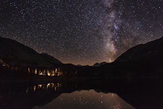 Stars fill the night sky, with a prominent Milky Way visible above a mountain range. Below, lights from a building or campsite illuminate trees near a reflective body of water, adding warmth to the scene.