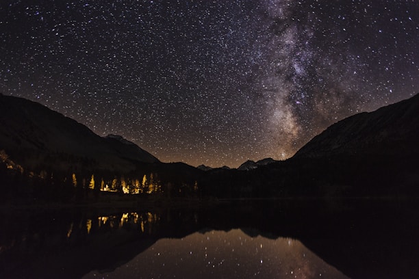 Stars fill the night sky, with a prominent Milky Way visible above a mountain range. Below, lights from a building or campsite illuminate trees near a reflective body of water, adding warmth to the scene.