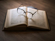 An open book resting on a wooden table with a pair of reading glasses beside it.