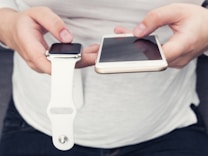 A person is holding a smartphone in one hand and a smartwatch with a white band in the other hand. The focus is on the electronic devices.