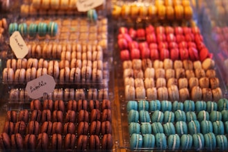 An array of colorful macarons neatly arranged in trays, featuring a variety of flavors indicated by small handwritten labels. The macarons display vibrant colors such as turquoise, pink, chocolate brown, and peach, with a delicate, glossy finish.