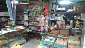 A cluttered small shop with shelves full of various items such as canned goods, packaged products, and household items. The counter is covered with miscellaneous objects including a fan, clock, and some papers. In the foreground, there are baskets containing eggs. The shop has a rustic and somewhat disorganized appearance.