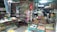 A cluttered small shop with shelves full of various items such as canned goods, packaged products, and household items. The counter is covered with miscellaneous objects including a fan, clock, and some papers. In the foreground, there are baskets containing eggs. The shop has a rustic and somewhat disorganized appearance.