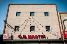 A beige building facade with four small windows displays the name 'C.H. MARTIN' in large red letters. Colorful triangular pennant flags are strung above, forming a festive appearance. Two 'FOR RENT' signs are visible on either side of the building.